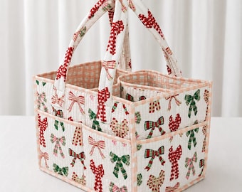 May include: A quilted fabric storage caddy featuring a white base with a repeating pattern of red and green bows. The caddy has two sturdy handles and a divided interior, with light pink and white gingham trim along the edges and handles.