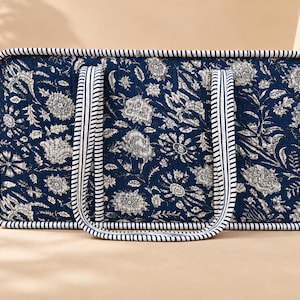 May include: A rectangular quilted tote bag featuring a dark blue fabric with a white floral pattern. The bag has two long handles and a thick border, both decorated with a navy blue and white striped design throughout the exterior.
