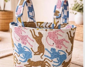 May include: A quilted fabric basket featuring a repeating pattern of stylized cheetahs in shades of pink, blue, and brown on a white background. The basket has two upright handles with a blue and white checkered pattern and stitched detailing.