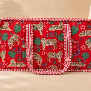 May include: A rectangular quilted tote bag in a vibrant red fabric featuring a repeating pattern of tigers, green monstera leaves, and small blue dots. The bag includes two matching red and white striped fabric handles and piped edges.