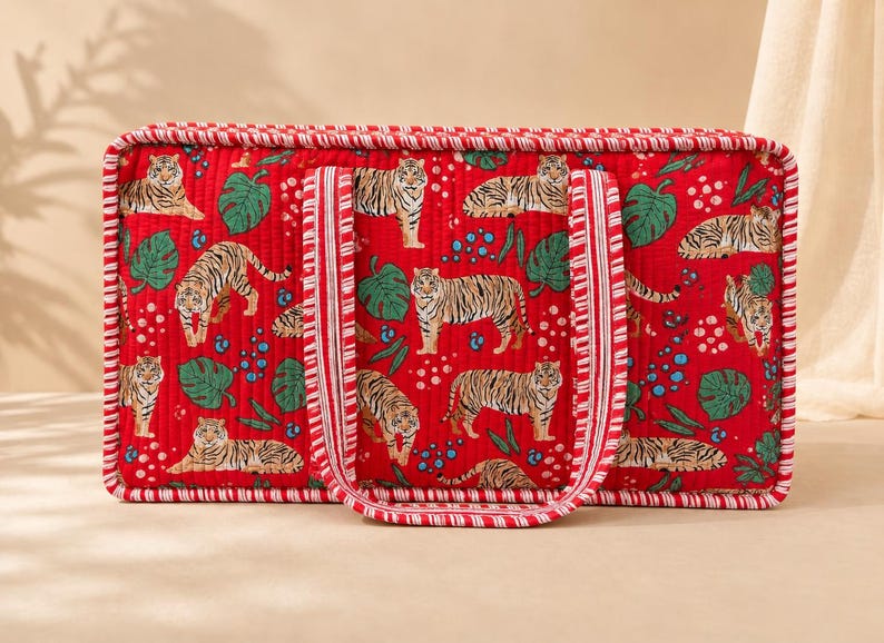 May include: A rectangular quilted tote bag in a vibrant red fabric featuring a repeating pattern of tigers, green monstera leaves, and small blue dots. The bag includes two matching red and white striped fabric handles and piped edges.
