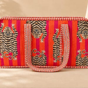 May include: A rectangular quilted tote bag featuring vertical stripes in bright orange and hot pink. The bag is decorated with a repeating pattern of black and beige tiger illustrations and has two matching fabric handles with striped trim.