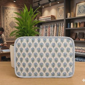 May include: A rectangular quilted fabric mat featuring a repeating pattern of blue and green floral motifs on an off-white background, finished with a light blue and white striped fabric border along the rounded edges.