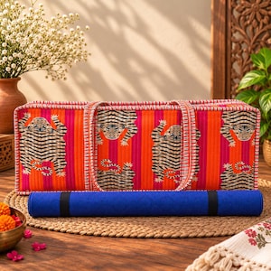 May include: A rectangular quilted fabric yoga mat bag featuring a vibrant orange and pink vertical striped pattern with repeating tiger illustrations. The bag has two matching fabric handles and sits behind a rolled solid blue yoga mat.