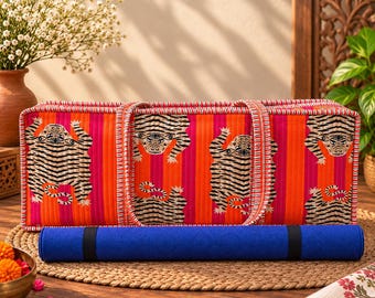 May include: A rectangular quilted fabric yoga mat bag featuring a vibrant orange and pink vertical striped pattern with repeating tiger illustrations. The bag has two matching fabric handles and sits behind a rolled solid blue yoga mat.