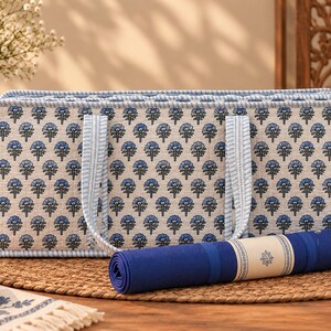 May include: A rectangular quilted storage bag featuring a blue and white floral block print pattern with light blue striped trim and handles, accompanied by a rolled solid dark blue yoga mat with a central cream-colored band.