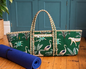 May include: A rectangular quilted fabric tote bag in dark green featuring a jungle print with zebras, leopards, and birds among palm leaves. The bag has two long striped handles and a front zippered pocket. A rolled blue yoga mat sits nearby.