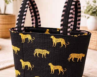 May include: A quilted fabric tote bag featuring a black base with a repeating pattern of yellow cheetahs. The bag has two sturdy handles with a black and pink striped design. The quilted texture is visible across the entire exterior surface.