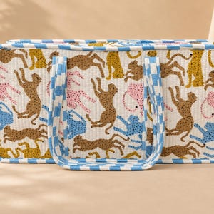 May include: A rectangular quilted tote bag featuring a repeating pattern of stylized leopards in shades of brown, yellow, pink, and blue on a white background. The bag has two handles and edges trimmed with a blue and white checkered fabric pattern.