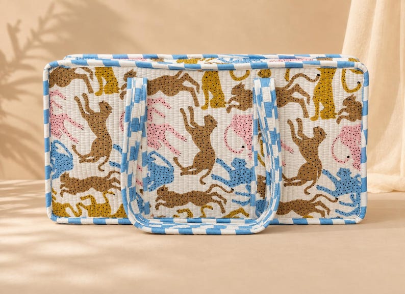 May include: A rectangular quilted tote bag featuring a repeating pattern of stylized leopards in shades of brown, yellow, pink, and blue on a white background. The bag has two handles and edges trimmed with a blue and white checkered fabric pattern.