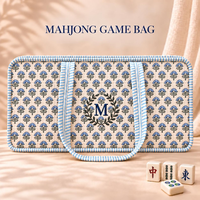 May include: A rectangular Mahjong game bag featuring a cream-colored fabric with a repeating blue floral pattern, blue and white striped trim, and a central dark blue letter M inside a laurel wreath. Three Mahjong tiles sit in front.