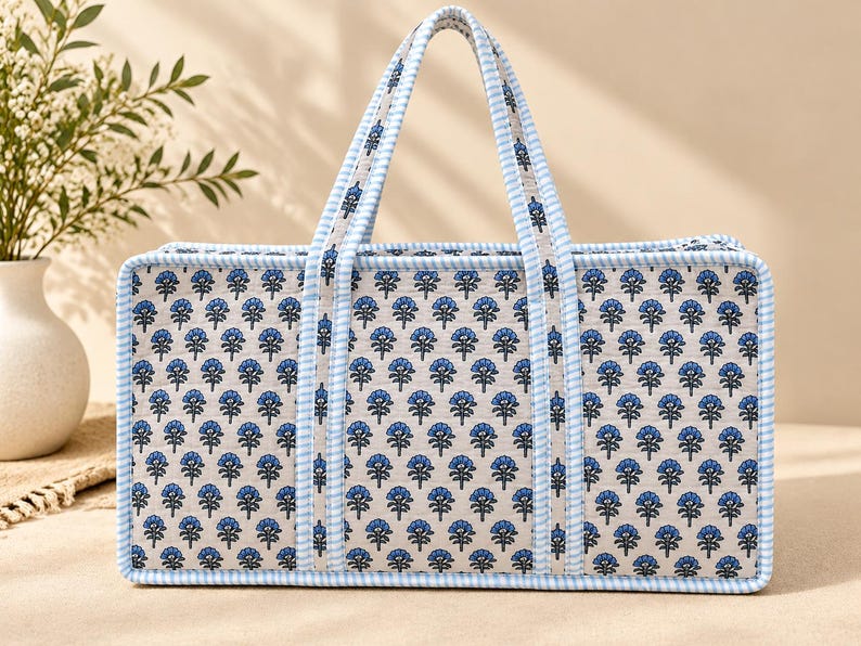 May include: A rectangular tote bag featuring a repeating blue floral pattern on a light beige background. The bag has two handles and edges trimmed with a light blue and white gingham check fabric, providing a structured appearance.