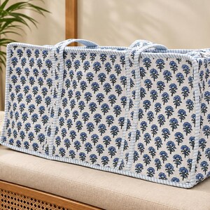 May include: A rectangular tote bag featuring a white fabric base with a repeating blue floral print. The bag has two matching fabric handles and is finished with light blue and white striped piping along all the edges and seams.