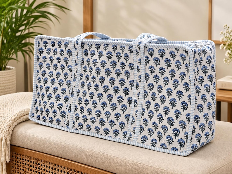 May include: A rectangular tote bag featuring a white fabric base with a repeating blue floral print. The bag has two matching fabric handles and is finished with light blue and white striped piping along all the edges and seams.