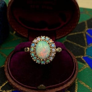 Könnte beinhalten: Ein goldener Ring mit einem ovalen Opal-Mittelstein, umgeben von kleineren Opalen. Der Ring ist in einem goldenen Band gefasst und wird in einer braunen, samtgefütterten Schmuckschatulle präsentiert.