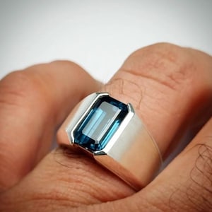 Op de afbeelding: Een zilveren ring met een rechthoekige, diepblauwe edelsteen. De ring heeft een brede, platte band en een bezel setting die de vorm van de edelsteen benadrukt. De edelsteen heeft een levendige, opvallende kleur.