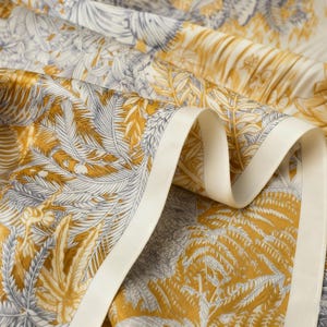 Puede incluir: Primer plano de una tela con estampado bot&aacute;nico en tonos dorados, grises y crema. El dise&ntilde;o presenta hojas y flores estilizadas. Se aprecia un borde de cinta color crema, que a&ntilde;ade un elemento decorativo al textil.