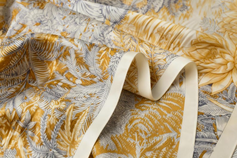 Puede incluir: Primer plano de una tela con estampado bot&aacute;nico en tonos dorados, grises y crema. El dise&ntilde;o presenta hojas y flores estilizadas. Se aprecia un borde de cinta color crema, que a&ntilde;ade un elemento decorativo al textil.