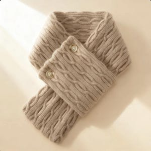 Könnte beinhalten: Beigefarbener Zopfstrickschal mit zwei Knopfverschlüssen. Der Schal besteht aus einem weichen, strukturierten Material und weist ein klassisches Zopfstrickmuster auf. Die Knöpfe sind hellbeige und passen zum Gesamtton des Schals.