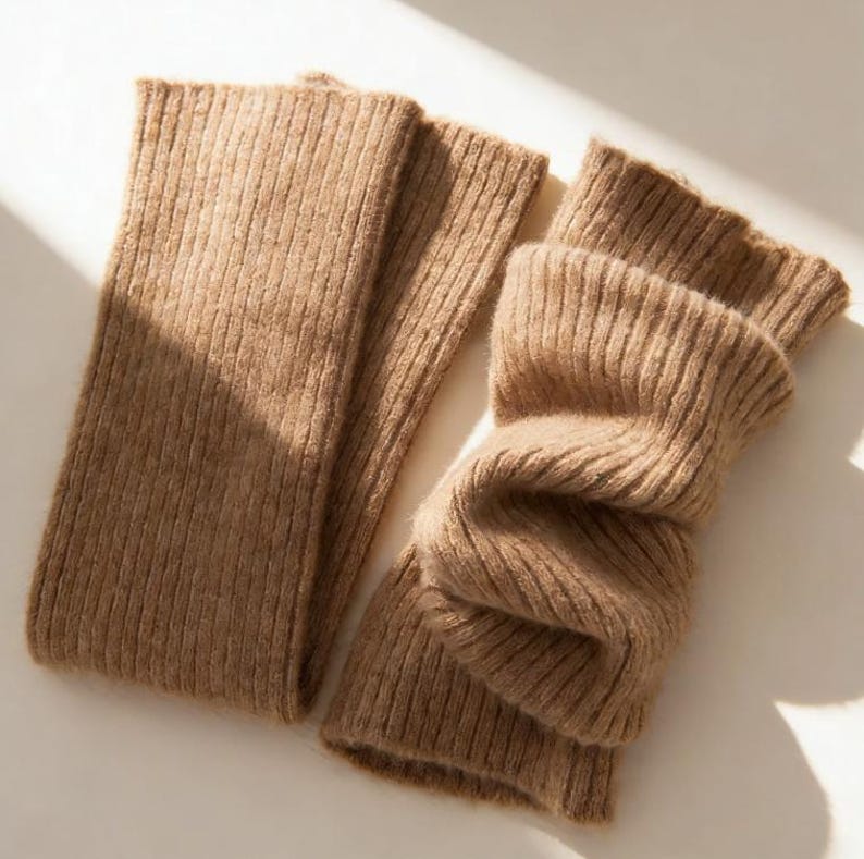 May include: Two tan-colored, ribbed-knit leg warmers. One is laid flat, while the other is folded, showing the texture of the soft material. The leg warmers are designed to keep legs warm and are a stylish accessory for colder weather.