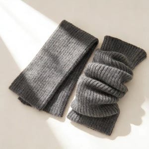 May include: A pair of gray knitted leg warmers. One is laid flat, while the other is folded and bunched. The ribbed texture is visible on both. The leg warmers are made of a soft, warm material.