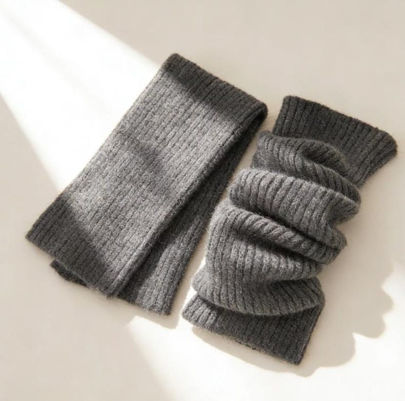 May include: A pair of gray knitted leg warmers. One is laid flat, while the other is folded and bunched. The ribbed texture is visible on both. The leg warmers are made of a soft, warm material.