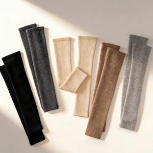 Peut inclure: Plusieurs jambières tricotées de couleurs neutres : noir, gris, beige et marron. Les jambières sont disposées sur une surface claire, mettant en valeur leur texture douce et leur palette de couleurs.