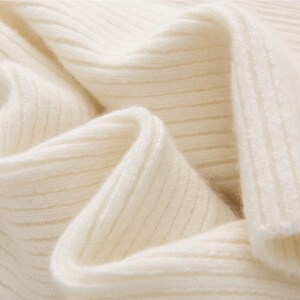 May include: Close-up of a cream-colored, ribbed knit fabric. The fabric is soft and appears to be folded, creating shadows and highlighting the texture. The knit pattern is visible, suggesting a sweater or other garment.