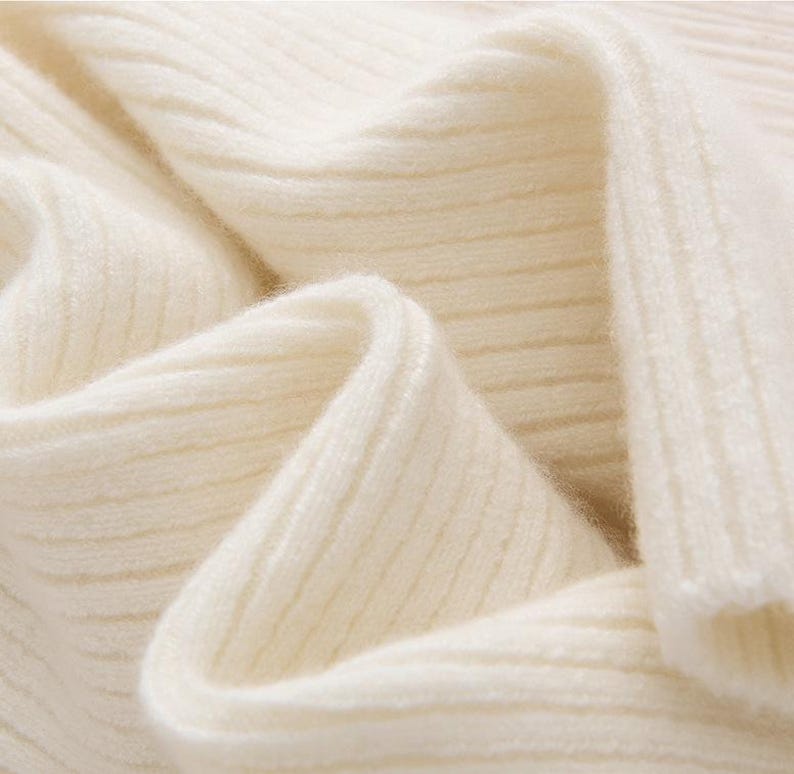 May include: Close-up of a cream-colored, ribbed knit fabric. The fabric is soft and appears to be folded, creating shadows and highlighting the texture. The knit pattern is visible, suggesting a sweater or other garment.