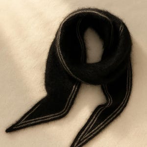 以下が含まれることがあります： 柔らかく、質感のある黒いスカーフ。スカーフの端には2本の細い平行なグレーのストライプがあり、暗い生地に微妙なコントラストを与えています。明るい色の表面にスカーフがかけられています。