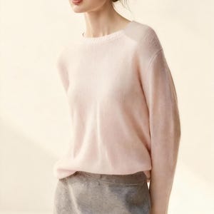 May include: A pale pink crew neck sweater with long sleeves. The sweater is worn with a grey skirt. The model is wearing gold earrings and has her hair up. The background is a light beige colour.