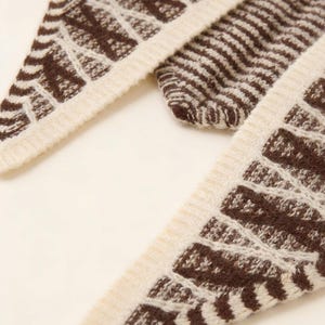 May include: Close-up of a knitted scarf with a geometric pattern. The scarf features a cream-colored border and a brown design. The pattern includes both solid stripes and openwork sections, creating a textured appearance. The scarf is made of a soft, warm material.