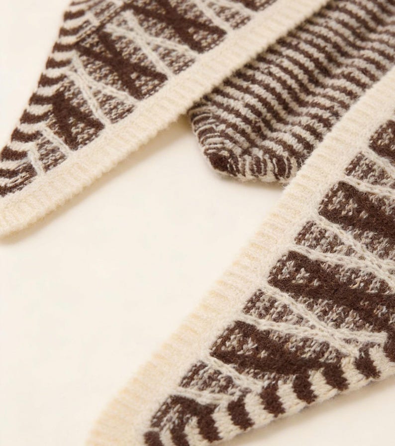 May include: Close-up of a knitted scarf with a geometric pattern. The scarf features a cream-colored border and a brown design. The pattern includes both solid stripes and openwork sections, creating a textured appearance. The scarf is made of a soft, warm material.
