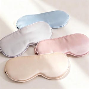 Könnte beinhalten: Fünf Schlafmasken aus Satin in Pastellfarben: Hellblau, Grau, Rosa und Beige. Die Masken haben eine geschwungene Form, um sich um die Augen anzupassen und sind so konzipiert, dass sie Licht blockieren, um den Schlaf zu verbessern. Die Masken sind aus einem weichen, glatten Material.