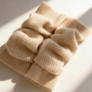 Könnte beinhalten: Ein gefalteter, beigefarbener Schal mit gerippter Strickstruktur. Der Schal besteht aus einem weichen Material und ist über sich selbst gefaltet, wodurch ein geschichteter Effekt entsteht. Der Schal liegt auf einer weißen Oberfläche.