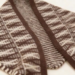 May include: Close-up of a brown and cream-colored cardigan. The knitwear features a geometric pattern with a dark brown border. The fabric appears soft and textured, suitable for cooler weather. The cardigan is folded, showing the collar and part of the sleeves.