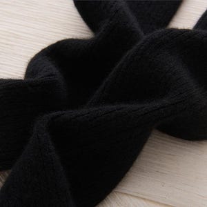 May include: Close-up of a black knitted fabric with a soft, textured appearance. The fabric is folded and draped, revealing the details of the knit. The image highlights the material's texture and color.
