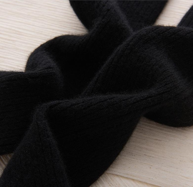 May include: Close-up of a black knitted fabric with a soft, textured appearance. The fabric is folded and draped, revealing the details of the knit. The image highlights the material's texture and color.