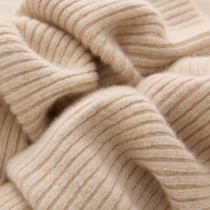 May include: Close-up of a beige ribbed knit fabric. The fabric is soft and appears to be made of a natural fiber. The texture is visible, with the ribbed pattern creating shadows and highlights.
