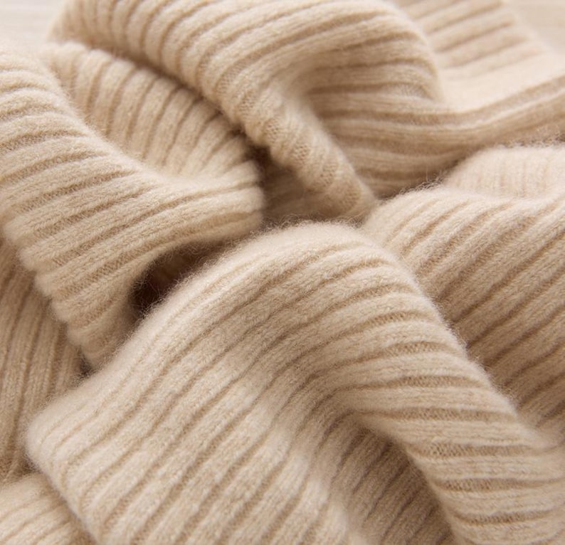 May include: Close-up of a beige ribbed knit fabric. The fabric is soft and appears to be made of a natural fiber. The texture is visible, with the ribbed pattern creating shadows and highlights.