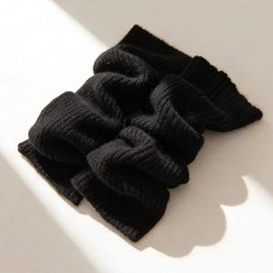 May include: A black, ribbed-knit scarf, folded and draped. The scarf is made of a soft material and has a textured appearance. The scarf is a solid black color and is displayed on a light-colored surface.