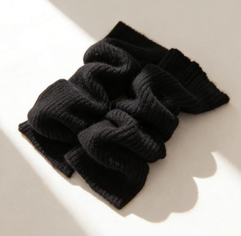 May include: A black, ribbed-knit scarf, folded and draped. The scarf is made of a soft material and has a textured appearance. The scarf is a solid black color and is displayed on a light-colored surface.