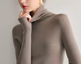 May include: A taupe turtleneck sweater with long sleeves. The ribbed knit fabric is visible. The model is wearing gold earrings. The sweater is a neutral color and appears to be made of a soft material.