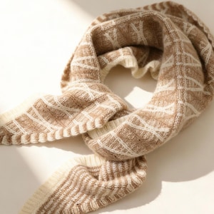 May include: A beige and cream-colored knitted scarf with a geometric pattern. The scarf is made of a soft, textured material and is draped in a circular shape. The design features a repeating diamond pattern.