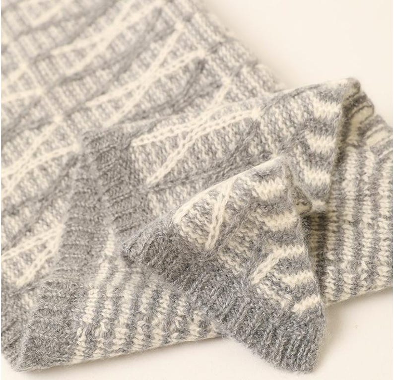 May include: Close-up of a folded gray and white knitted fabric. The knitwear features a repeating geometric pattern in a light gray and off-white color scheme. The texture of the fabric is visible, suggesting a soft and warm material.
