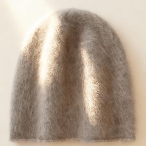 Könnte beinhalten: Eine hellgraue Beanie-Mütze mit weicher, flauschiger Textur. Die Mütze besteht aus einem flauschigen Material und hat eine einfache, abgerundete Form. Die Mütze hat eine neutrale Farbe und ist für verschiedene Outfits geeignet.