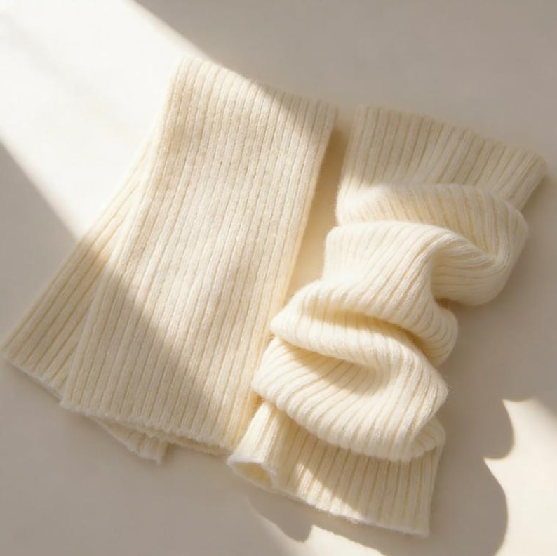 May include: A cream-colored, ribbed knit scarf, folded and draped. The scarf is made of a soft, textured material. The design features a simple, classic style, suitable for cold weather. The scarf is folded over itself, creating a layered effect.