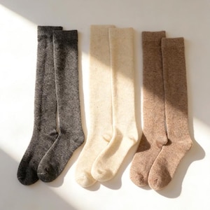 Könnte beinhalten: Drei Paar kniehohe Socken in einer neutralen Farbpalette. Die Socken werden nebeneinander präsentiert, mit Paaren in Anthrazitgrau, Creme und Hellbraun. Die Socken scheinen aus einem weichen, strukturierten Material zu sein.
