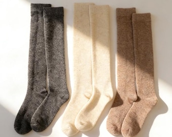 May include: Three pairs of knee-high socks in a neutral color palette. The socks are displayed side-by-side, with pairs in charcoal gray, cream, and light brown. The socks appear to be made of a soft, textured material.