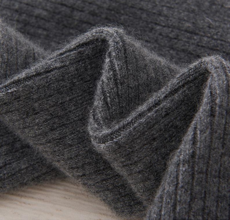 May include: Close-up of a section of dark gray ribbed knit fabric. The fabric is folded, showing the texture and the way the light plays across the surface. The knit has a soft, slightly fuzzy appearance.